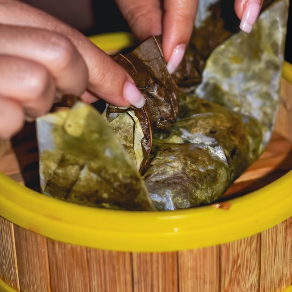 Hands with pink nails arrange leafy greens and ingredients in a bright yellow bowl on a wooden table.