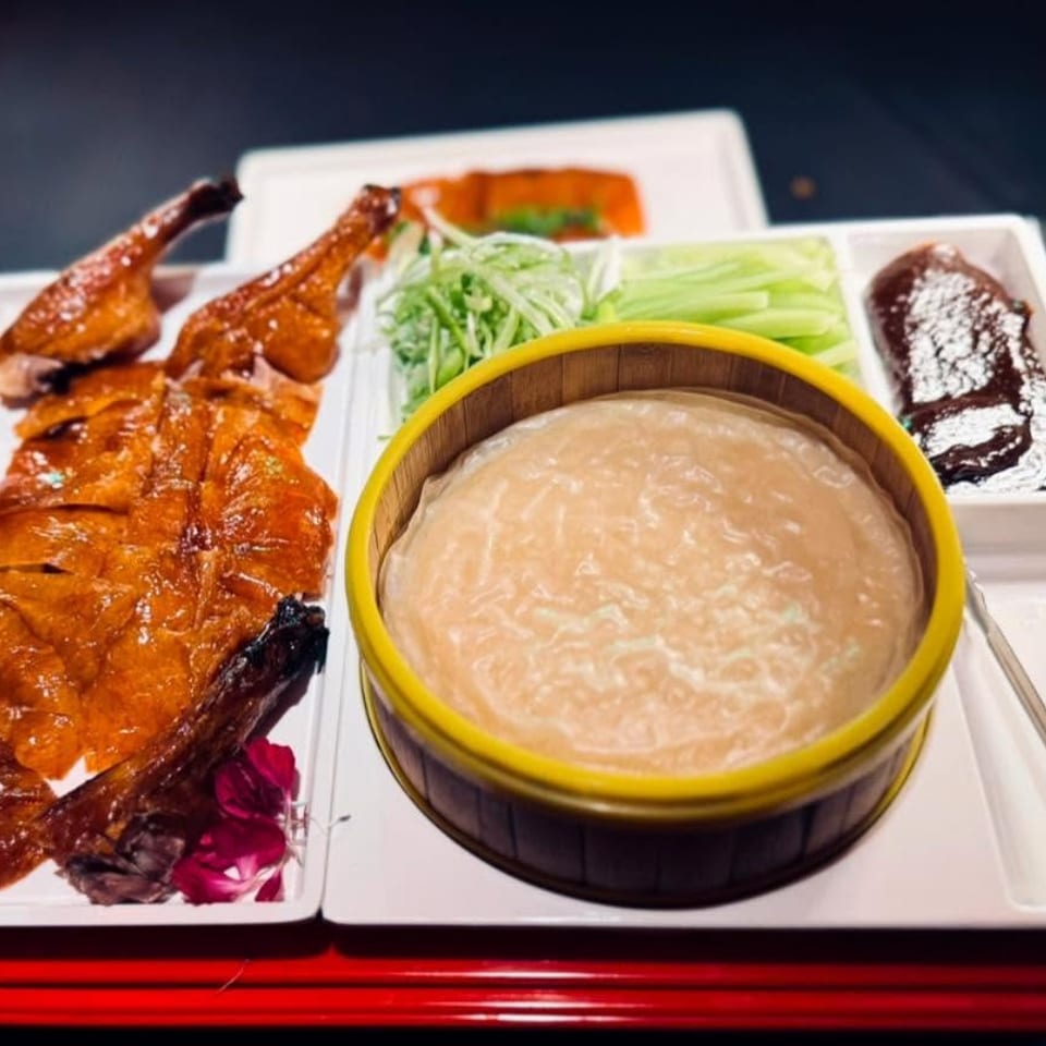 Plated Chinese roast duck with pancakes, sliced scallions, cucumber, and dark hoisin sauce beside a bamboo steamer.
