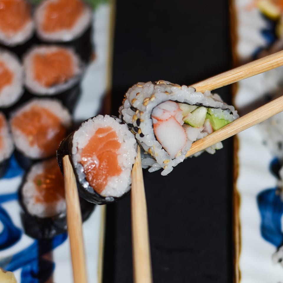 Chopsticks hold assorted sushi rolls with salmon, shrimp, avocado, and cucumber over a platter of sushi.