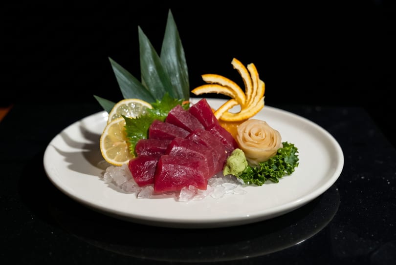 Tuna sashimi on ice with lemon, ginger, wasabi, and orange peel on a white plate.