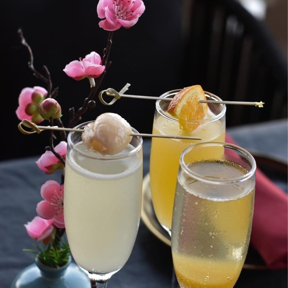 Three sparkling cocktails garnished with fruit skewers sit beside pink blossoms on a restaurant table.