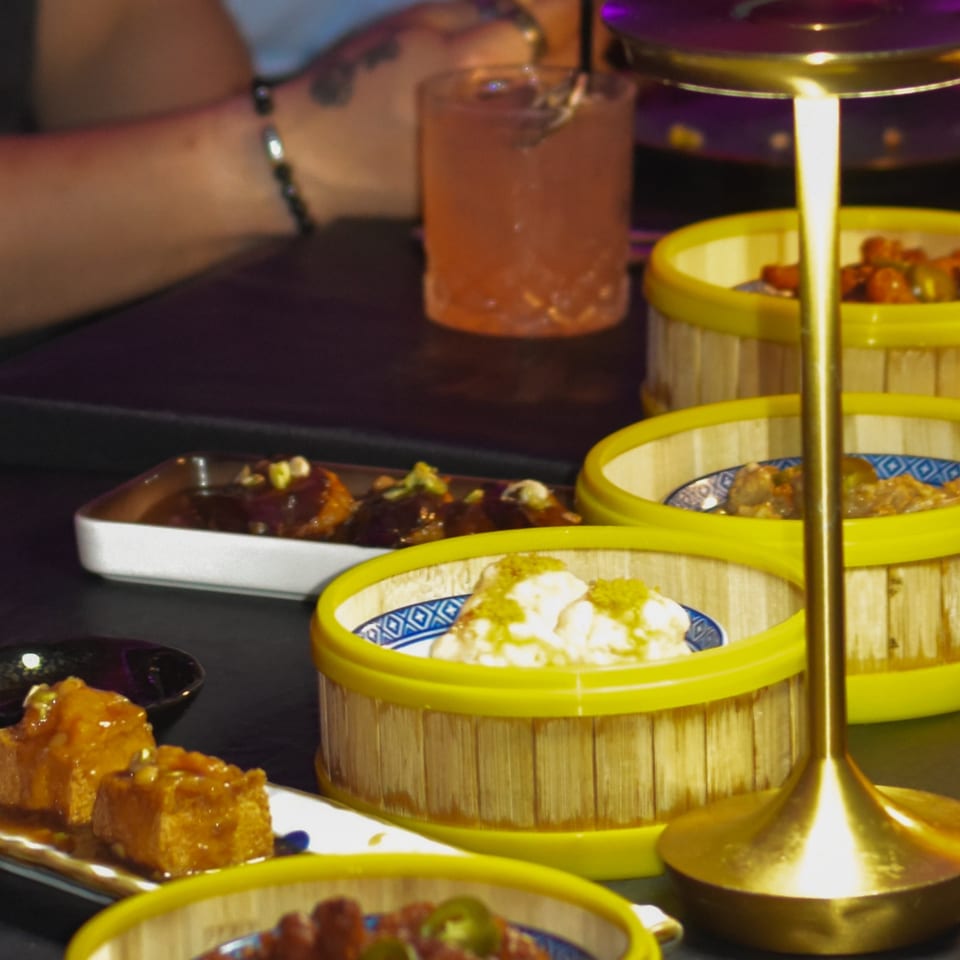 Colorful dim sum baskets filled with assorted dishes surround a cocktail on a restaurant table, with diners in the background.
