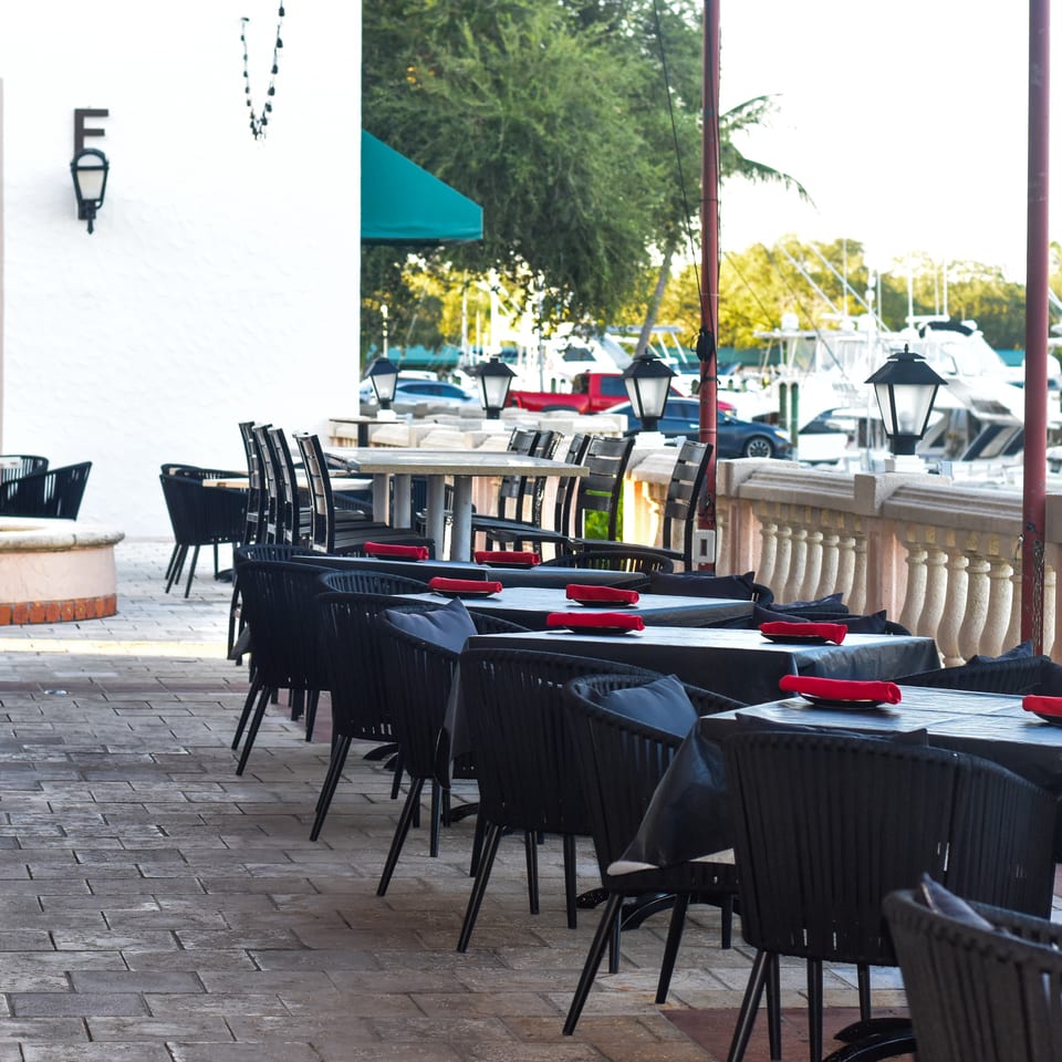 Outdoor restaurant patio with black tables, wicker chairs, red napkins, decorative lanterns, and boats visible along the waterfront.