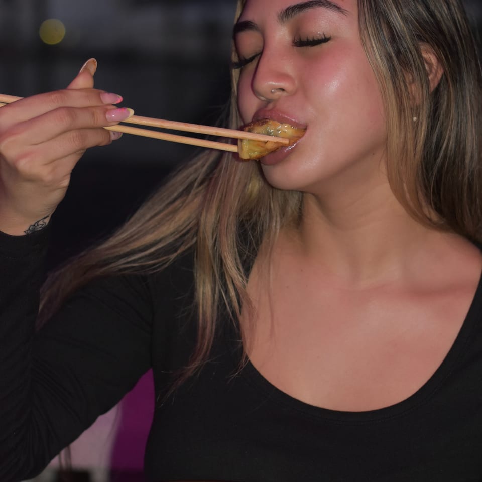 Woman eating a golden fried appetizer with chopsticks while holding a black plate in a dimly lit restaurant.