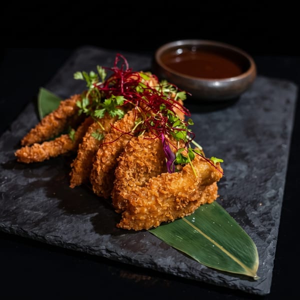 Crispy fried chicken or fish pieces, garnished with herbs, served on a dark slate with dipping sauce.