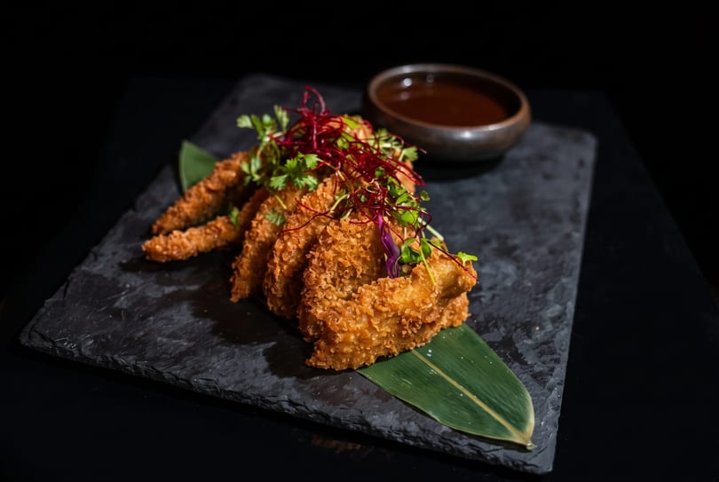 Crispy fried chicken or fish pieces, garnished with herbs, served on a dark slate with dipping sauce.