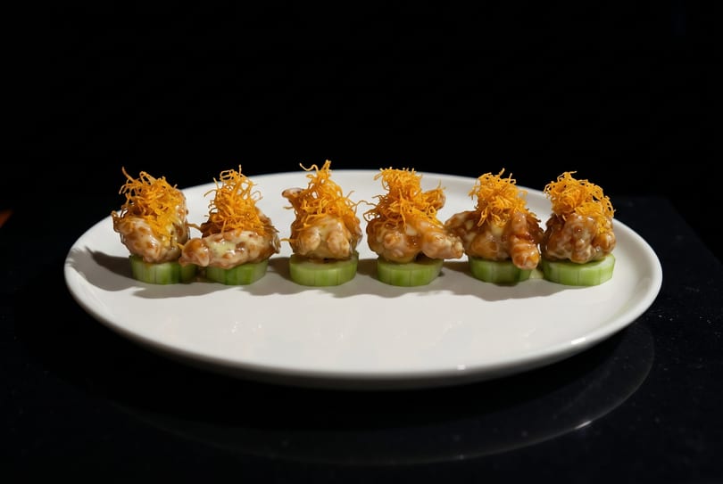 Six pieces of crispy shrimp with sauce and fried noodles on cucumber slices, on a white plate.