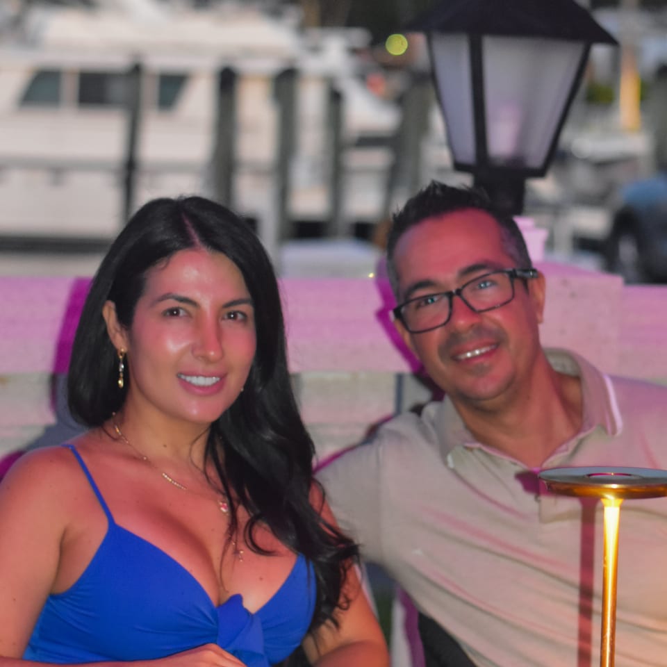 Two smiling diners sit at a restaurant table with dim sum, plated appetizers, menus, and warm decorative lighting.