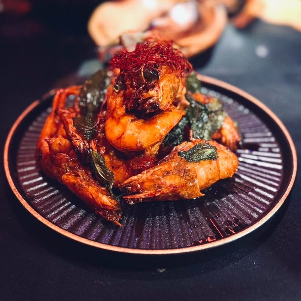Spicy grilled prawns garnished with crispy chili and leafy herbs on a black ridged plate.