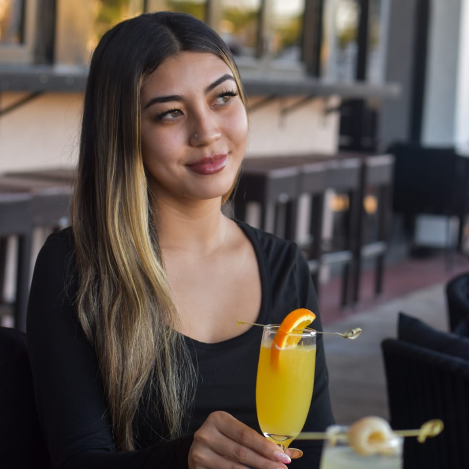 Woman in a restaurant holding an orange cocktail garnished with citrus, with another drink and folded napkin on the table.