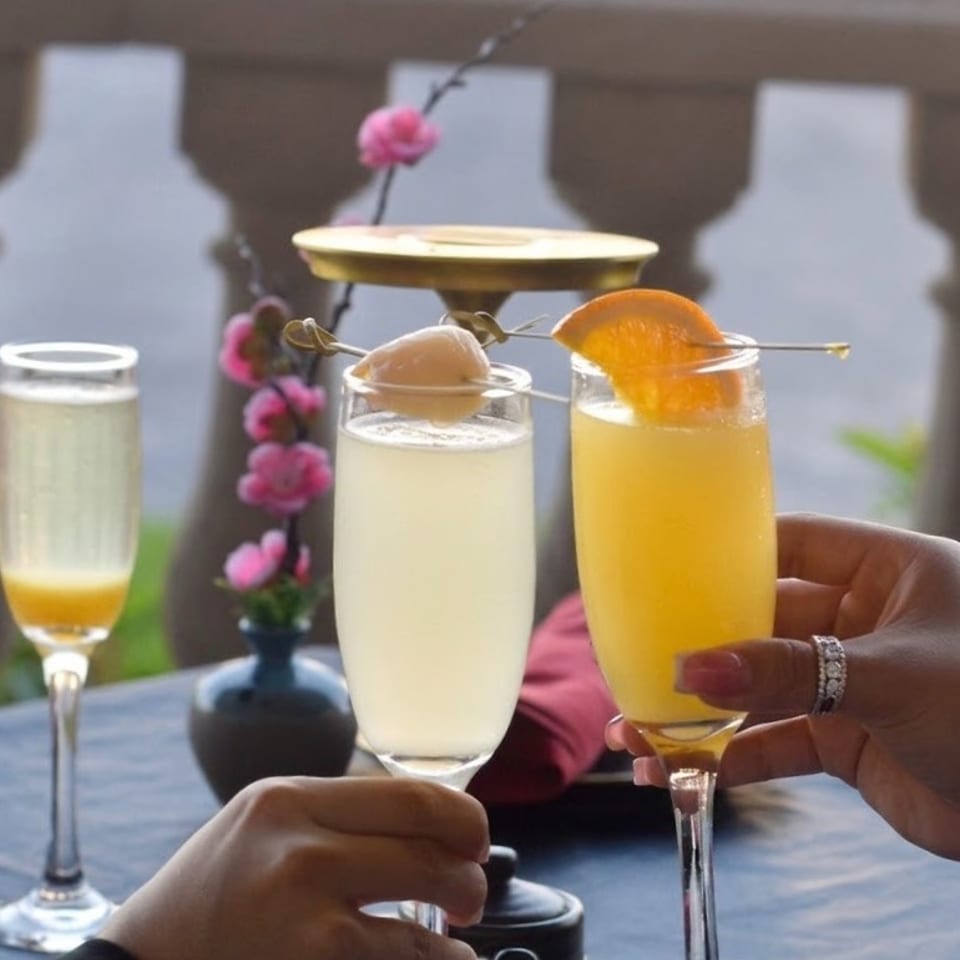 Hands raise two champagne flutes filled with pale citrus cocktails, garnished with fruit, beside a flowering plant outdoors.