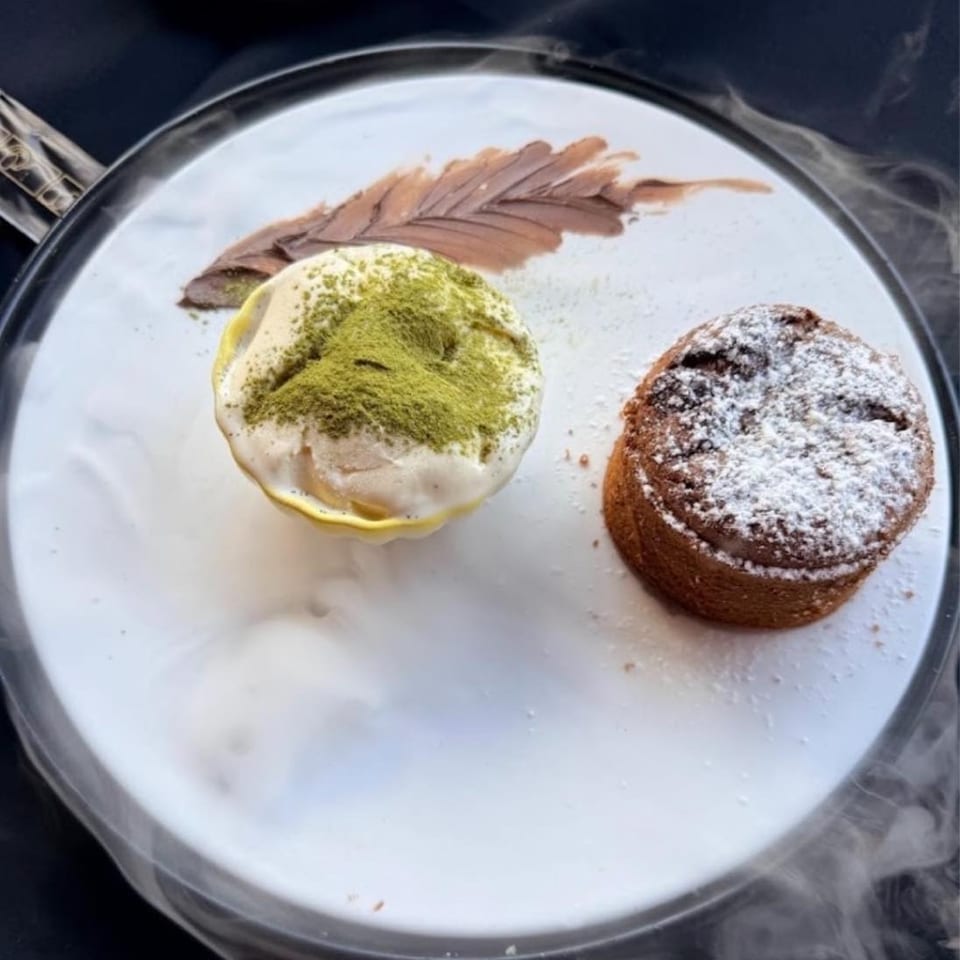 Dessert plate featuring matcha-dusted cream, a powdered sugar pastry, and chocolate sauce garnish on a white plate.
